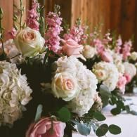 Pastel pink and white floral arrangements lined along a wooden table in a softly lit setting