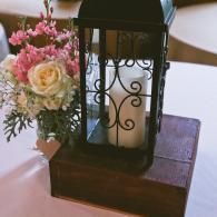 Black lantern centerpiece on a wooden box beside a pink-and-white floral arrangement on a table.