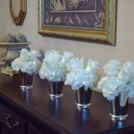 Four silver vases with white flowers arranged on a dark wooden dresser under framed wall art