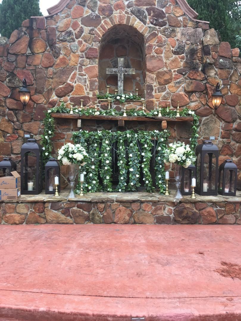 Stone grotto with a cross, greenery, lanterns, and candles on a red patio floor