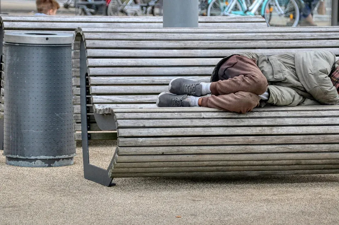 Person curled up sleeping on a wooden bench outdoors, next to a trash can.