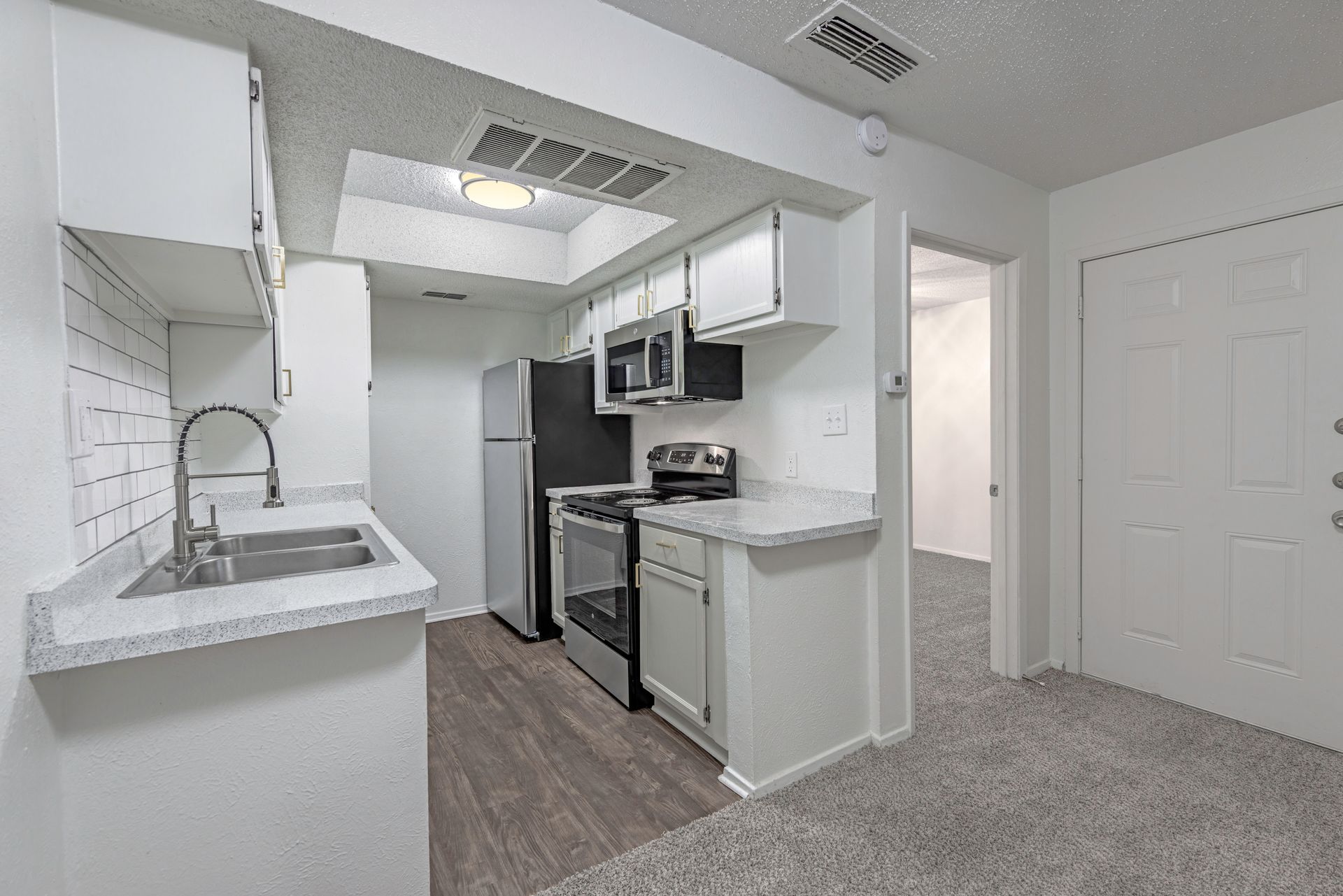White kitchen with stainless steel appliances, grey countertops, and a door to a carpeted area.