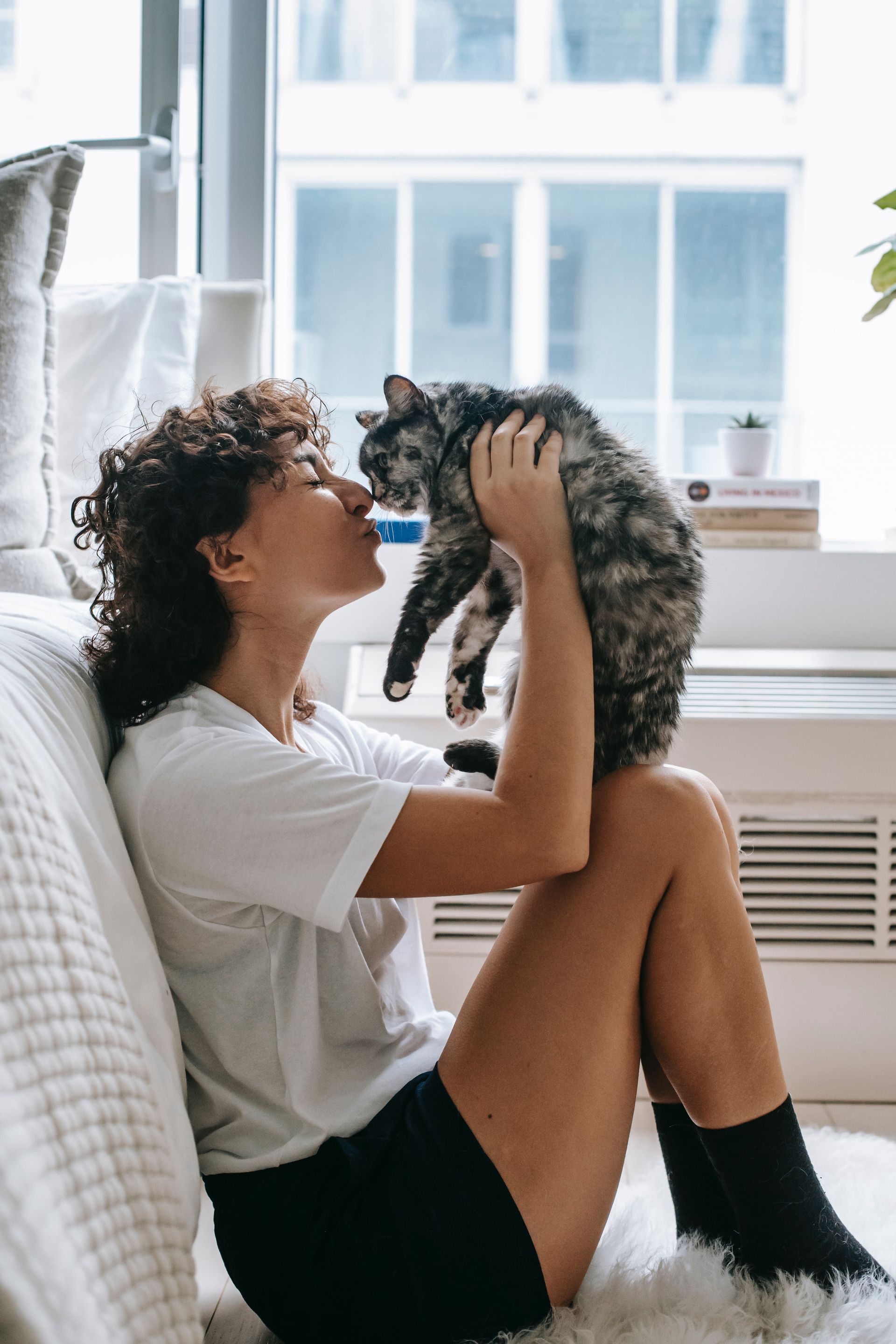 Woman kissing a gray tabby cat indoors, near a window. Both are smiling.