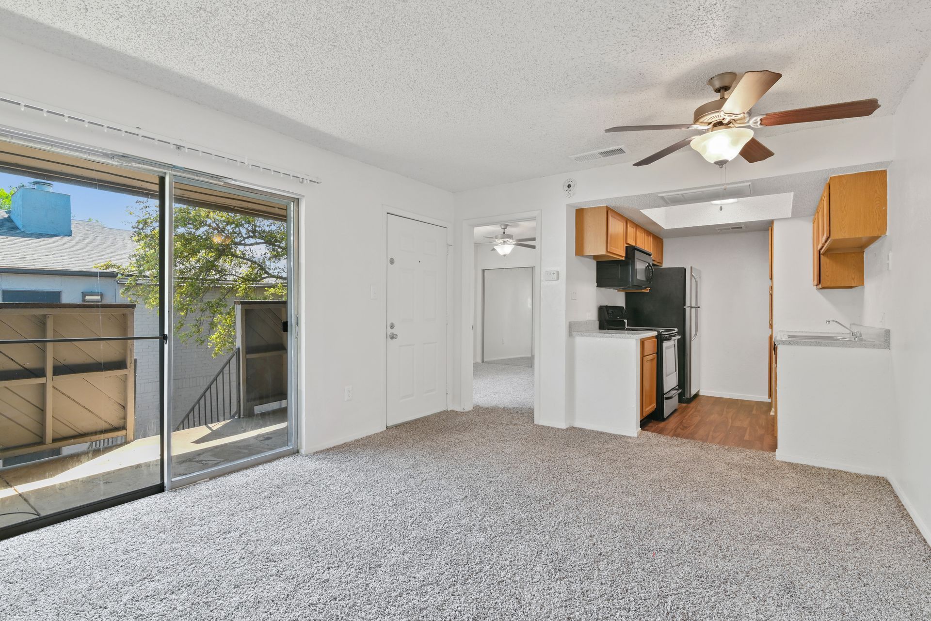 A living room with sliding glass doors, a kitchen, light-colored carpet, and a ceiling fan.