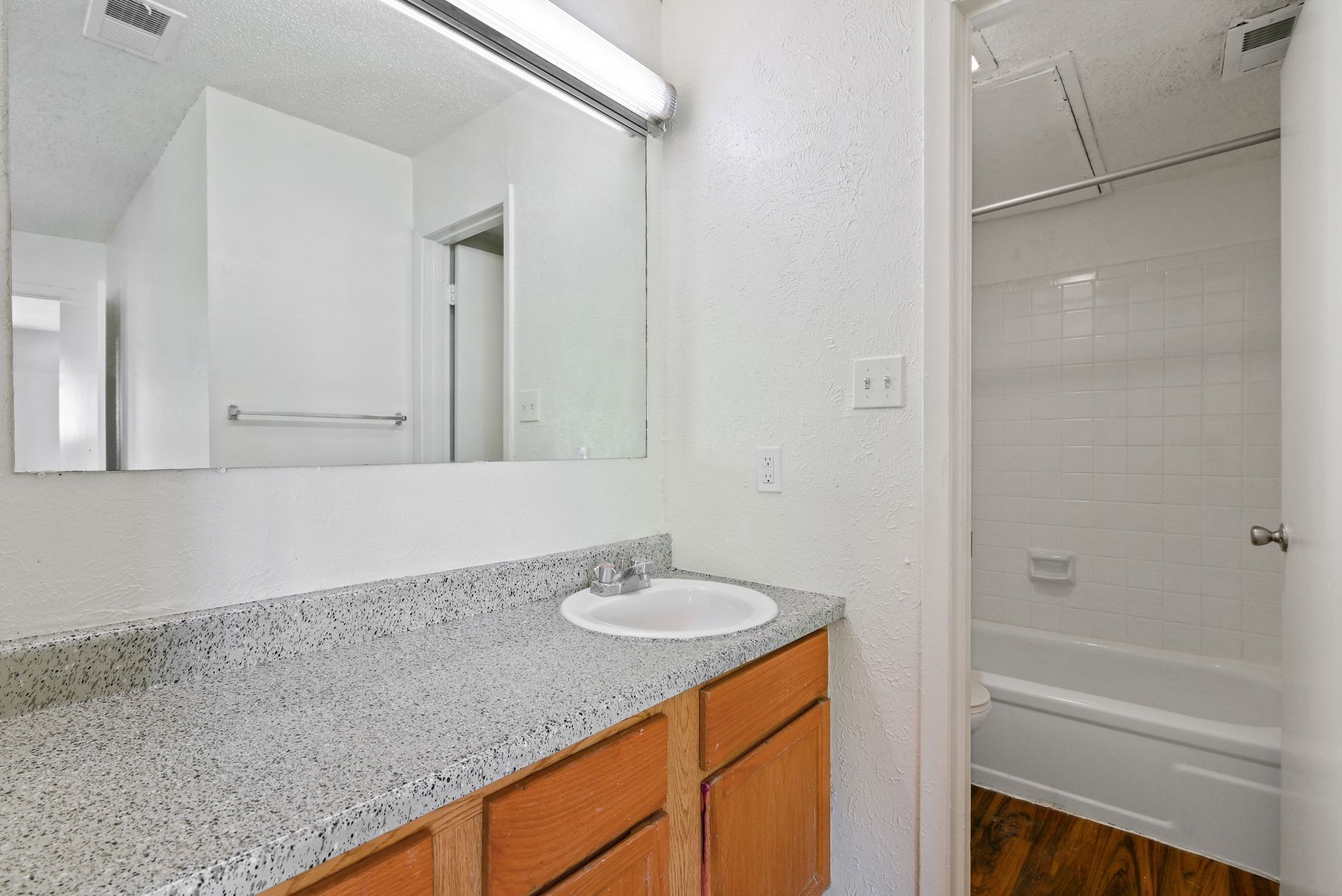 Bathroom with a vanity, mirror, and bathtub. The countertop is gray and white, the cabinets are wood.