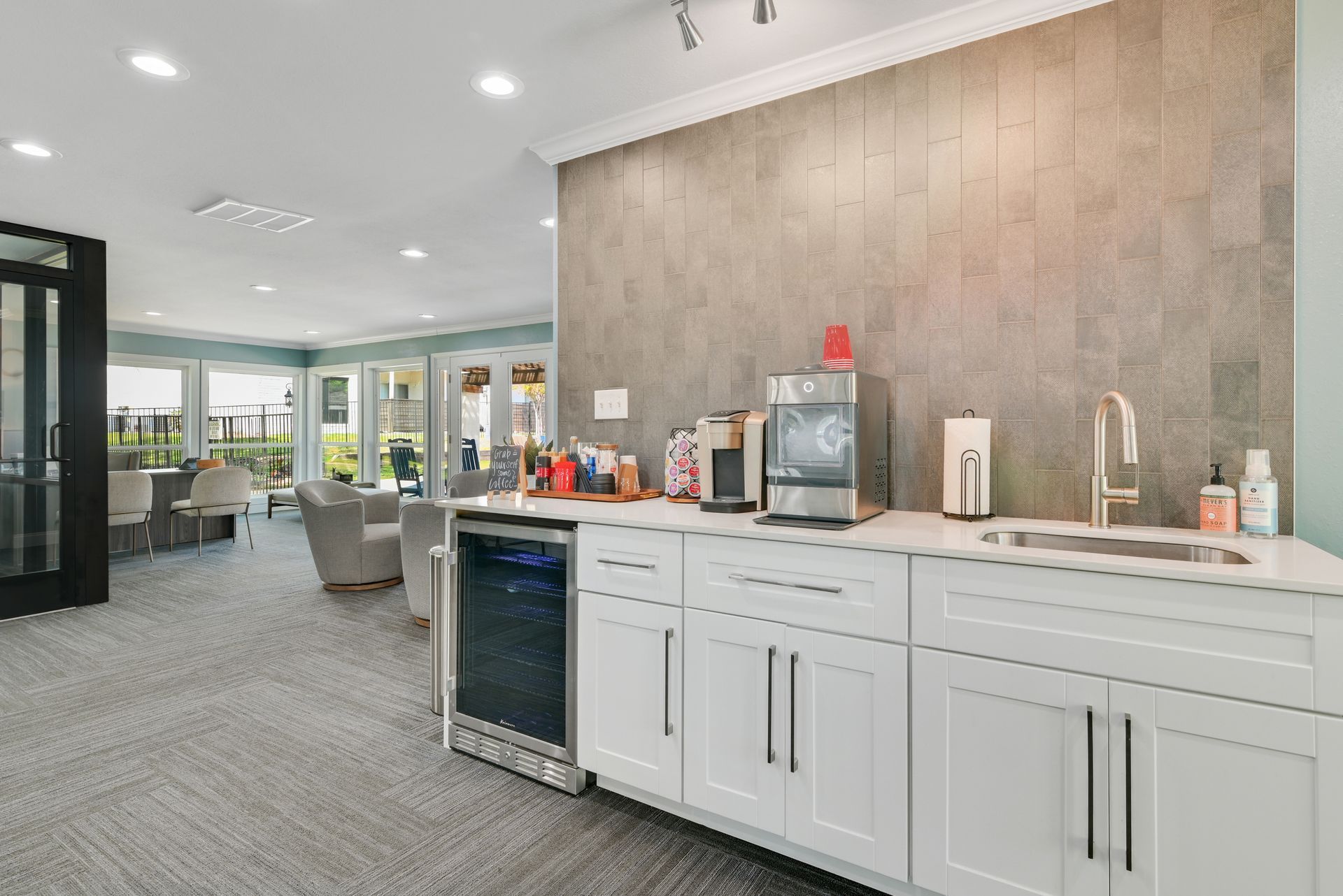 A modern lounge with a beverage bar and seating area, light gray walls, and large windows.