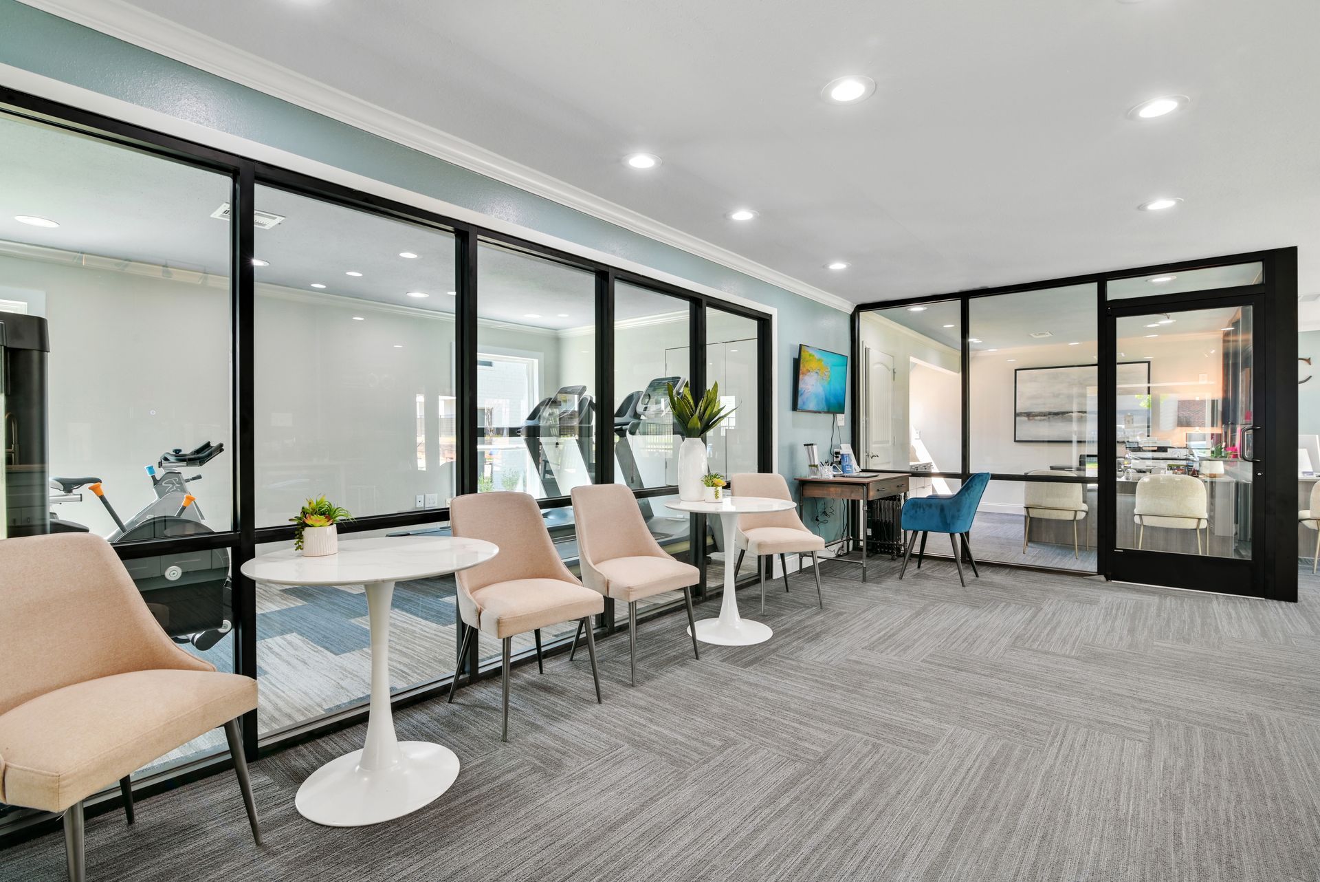 Modern waiting area with glass walls, light-colored chairs, small tables, and a glimpse of a fitness room.