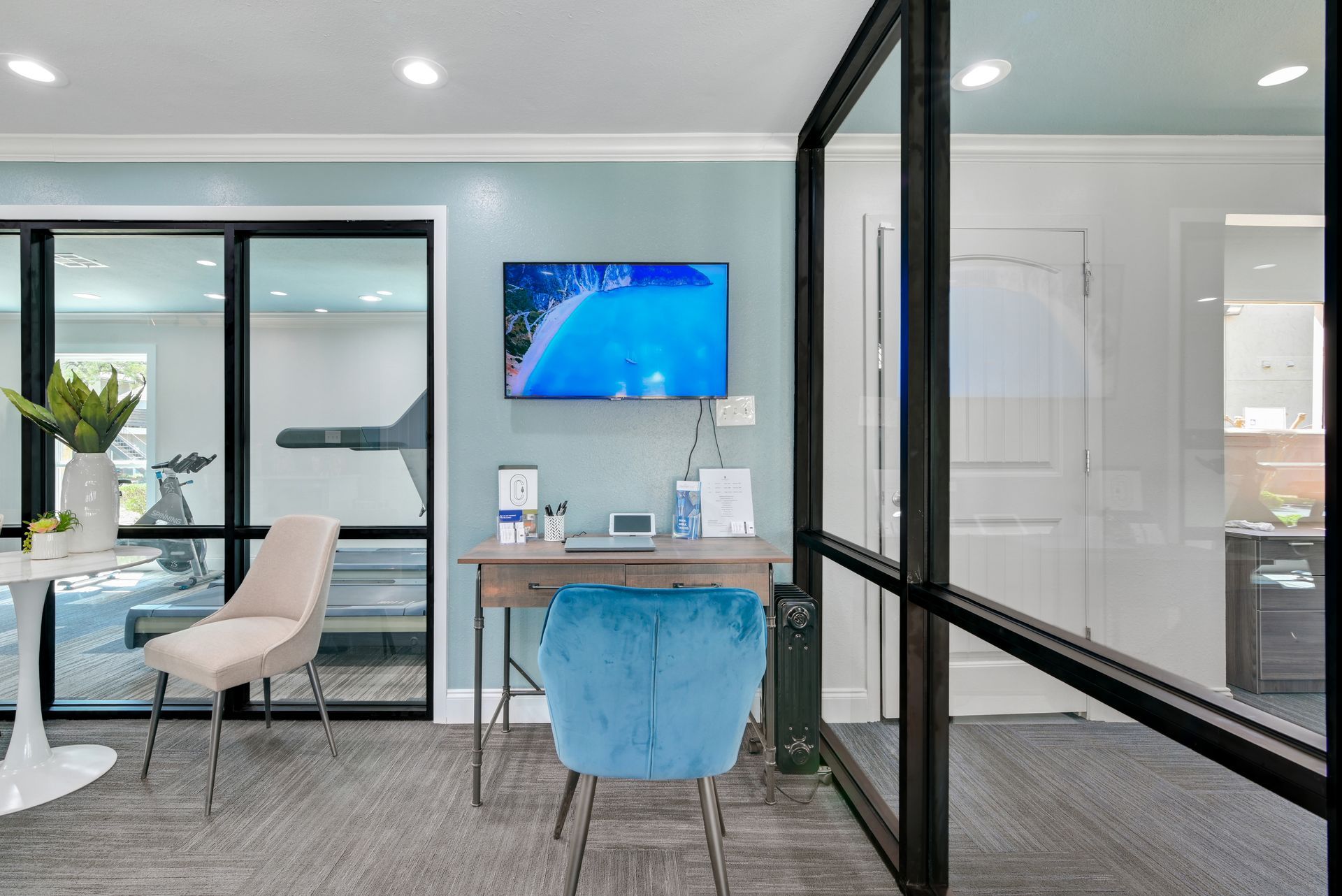 Office space with glass walls, desk, blue chair, TV, and a treadmill in the background.