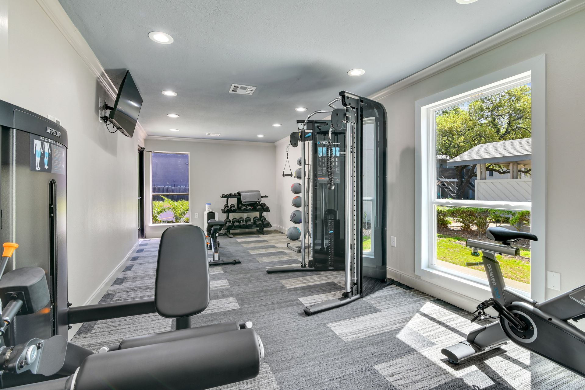 Gym interior with exercise equipment: weight machines, bike, and weights. Window with outside view.