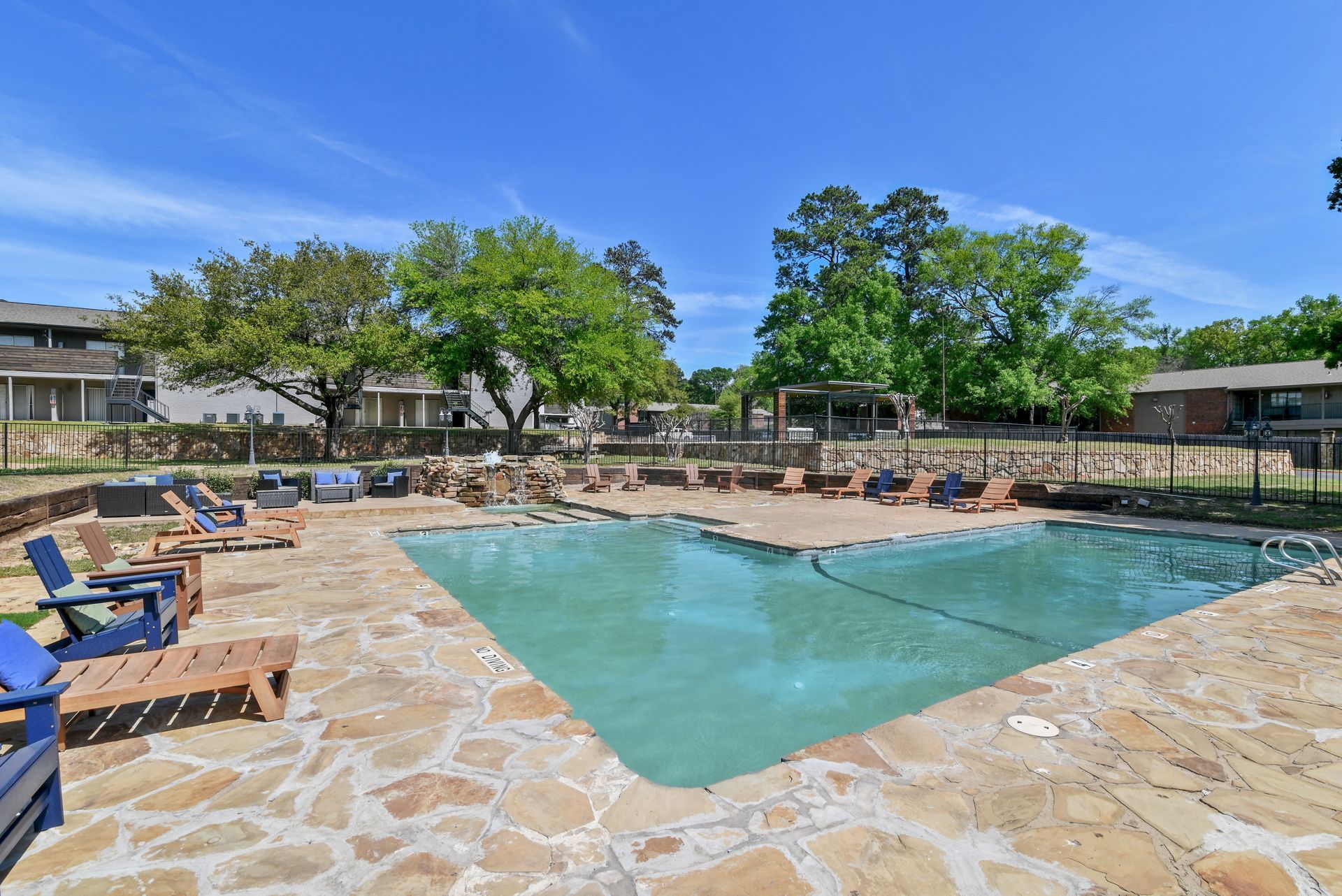 Pool area with lounge chairs, surrounded by trees and buildings under a blue sky.