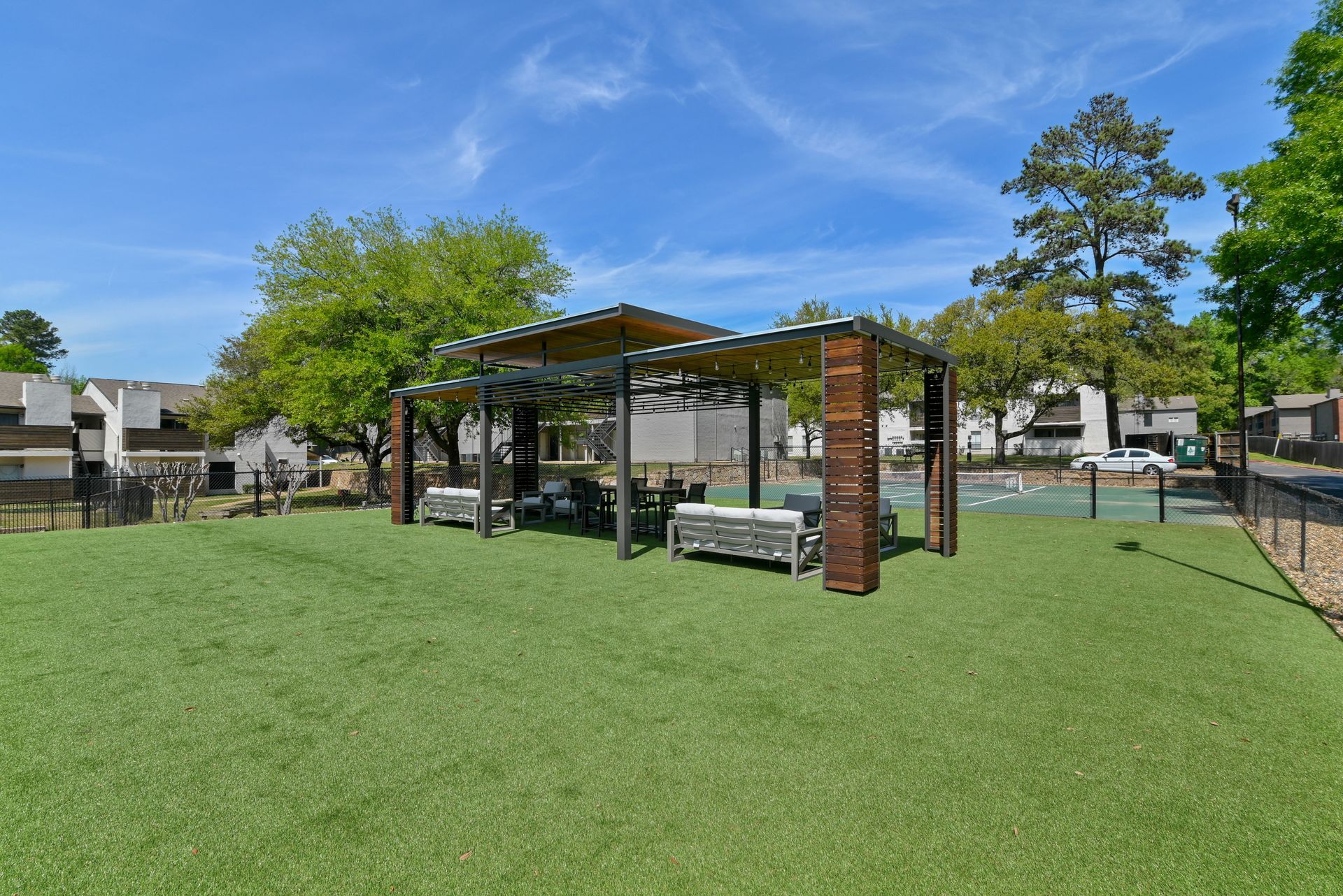 Outdoor seating area with pergola, on artificial grass, surrounded by apartment buildings, trees, and blue sky.