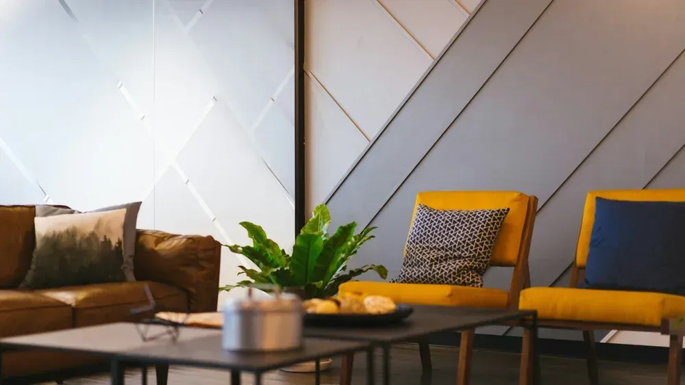 Modern lounge with brown sofa, yellow chairs, coffee table, and potted plant against geometric wall panels