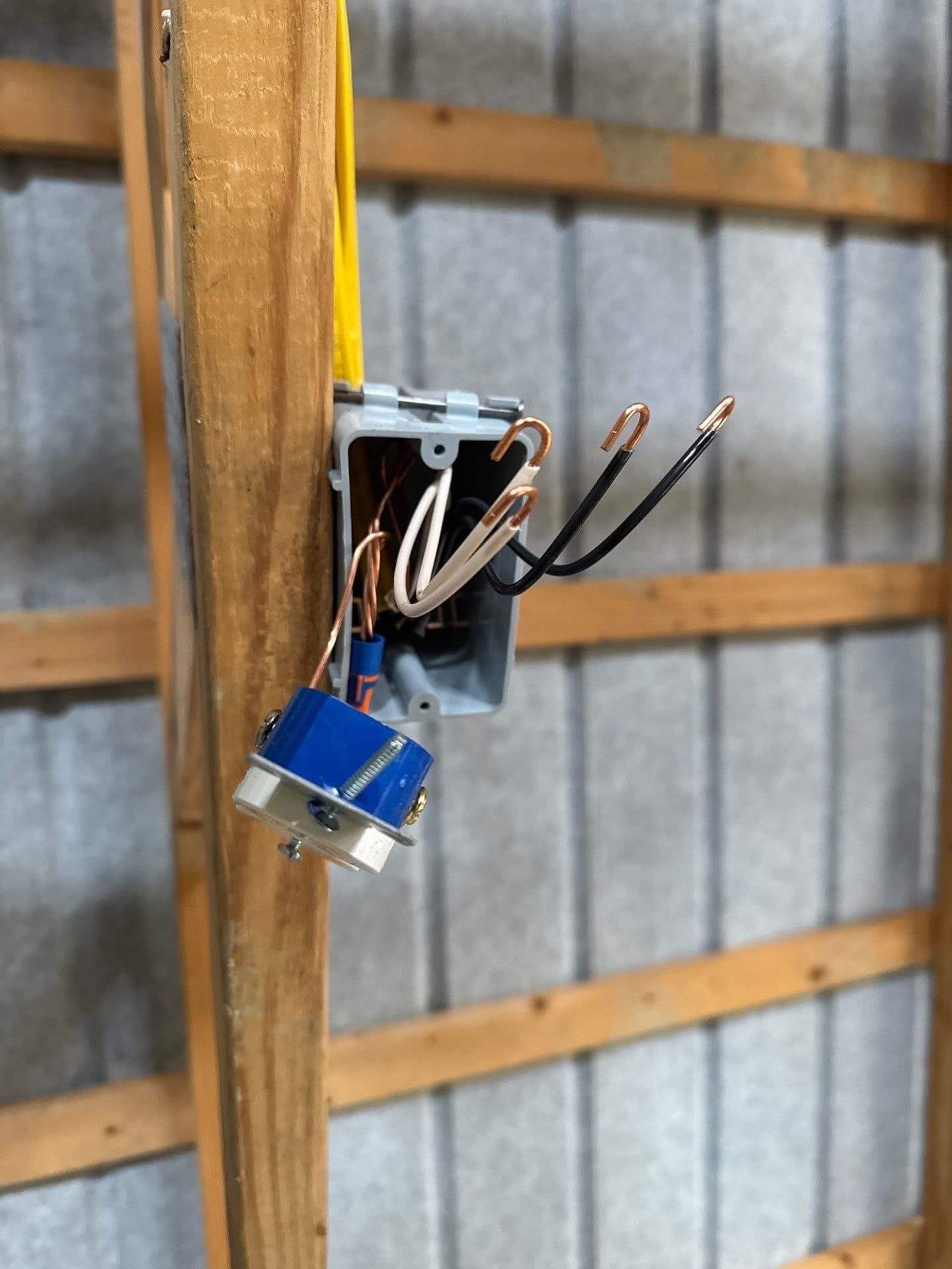 A gray electrical box attached to a wooden stud with exposed white, black, and copper wiring and a blue wall switch.