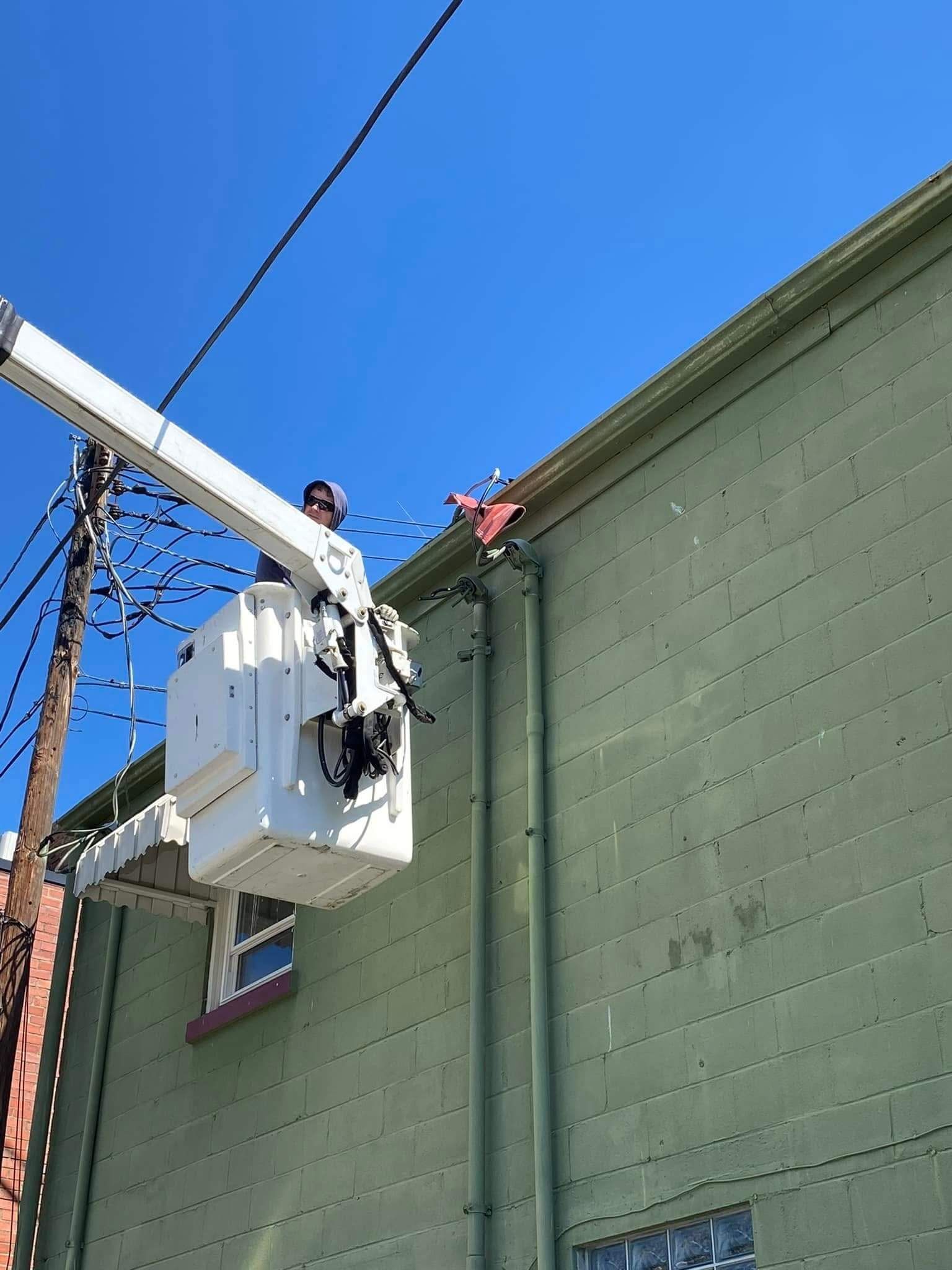 A worker in a white bucket truck boom reaches toward an electrical line attached to a light green brick building.