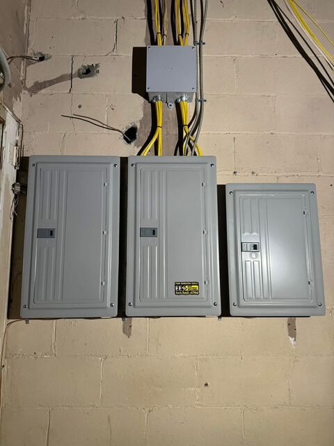 Three gray electrical breaker panels mounted on a light-colored cinder block wall with yellow conduit wiring above.