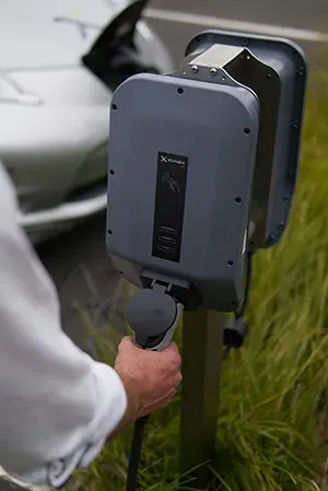 A person holding an electric vehicle charging connector in front of a gray charging station and a parked white car.