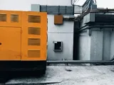 A yellow industrial generator sits on a concrete surface next to a white wall with conduit pipes and electrical boxes.