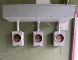 Three grey electrical meter sockets mounted on a green cinder block wall beneath a large rectangular electrical box.