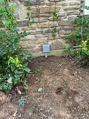 A gray electrical junction box mounted on a stone wall above bare soil, surrounded by greenery.