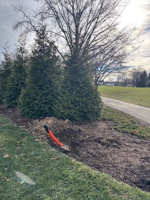 A person wearing work clothes and gloves reaches into a muddy, narrow trench containing cables.