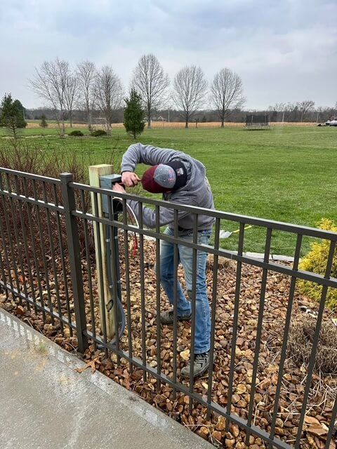 A person in a gray hoodie and jeans works on electrical wiring attached to a wooden fence post in a grassy yard.