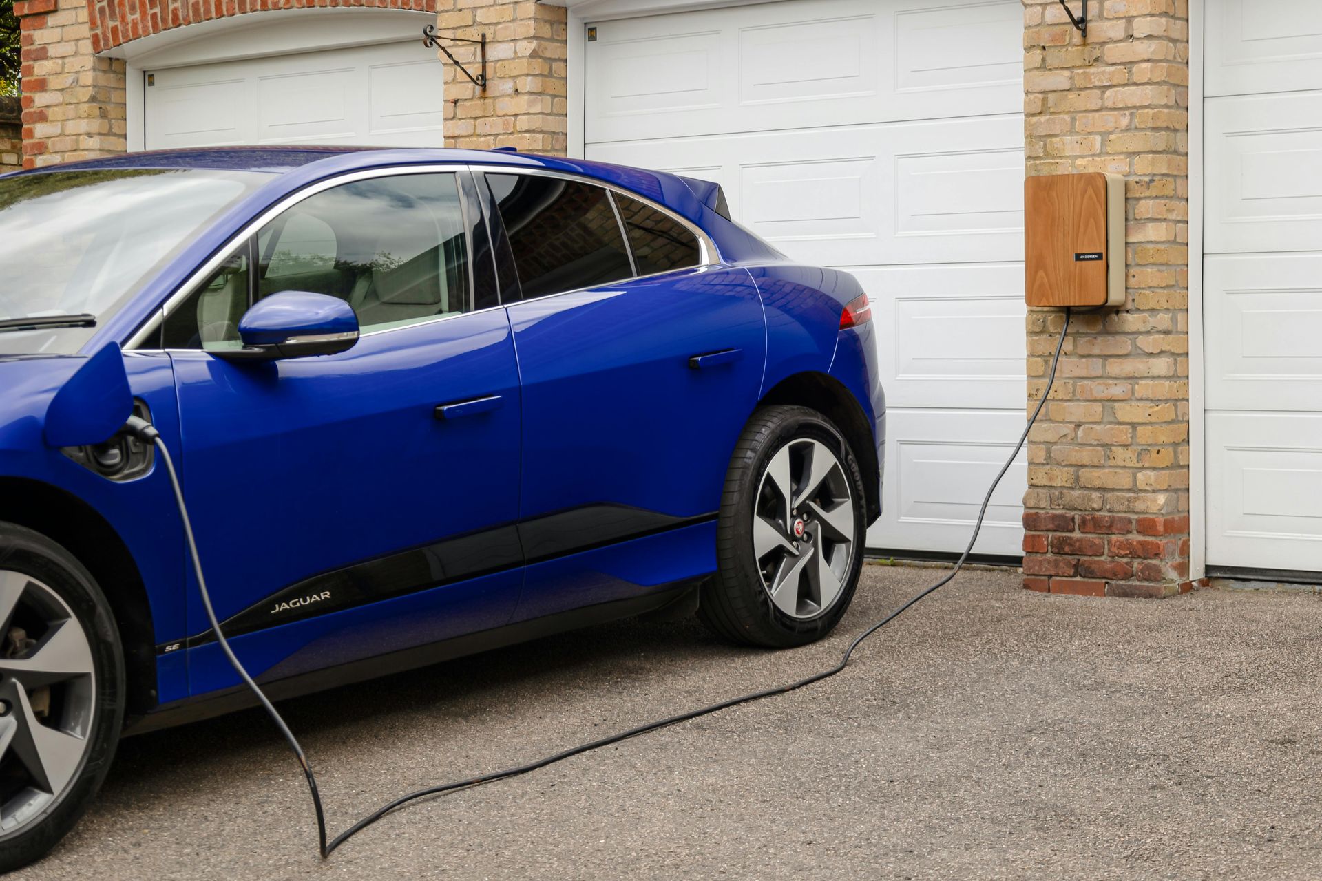 A blue electric car parked in a driveway, plugged into a wall-mounted charging station on a brick garage.