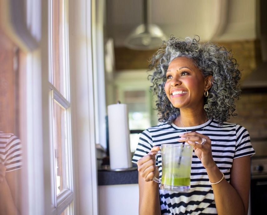 Smiling woman with grey curly hair, holding a green drink by a window in a kitchen, wearing a striped shirt.