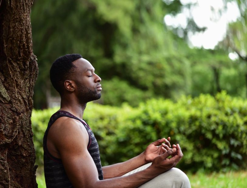A Black man meditates in a park, leaning against a tree, eyes closed, hands in prayer pose.
