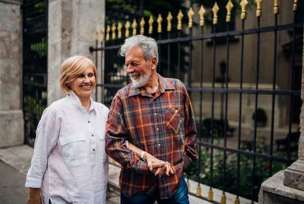 Elderly couple walking, smiling, arm-in-arm, near a decorative iron gate and stone structure.