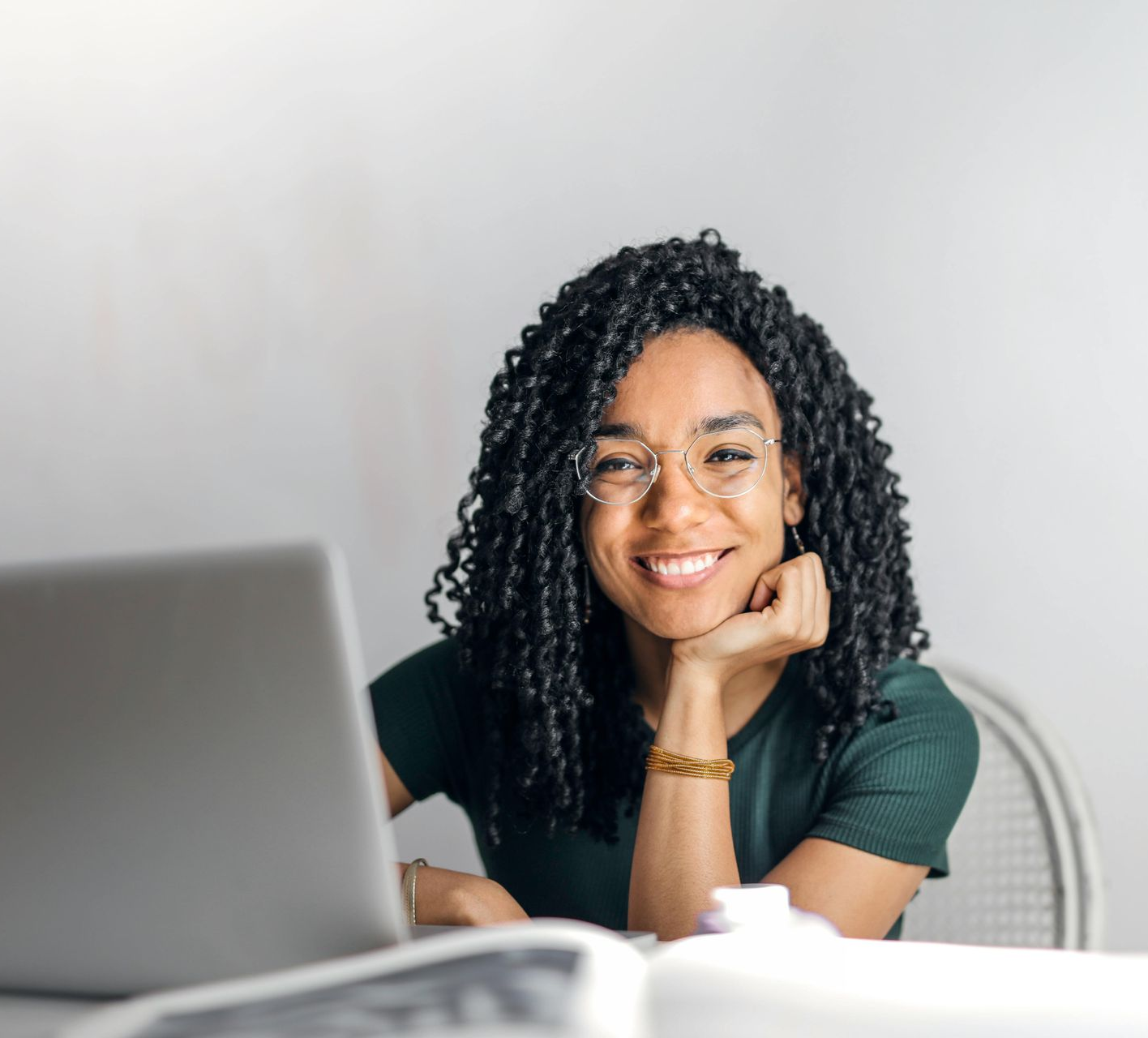Woman with glasses and black braided hair smiles while seated at a table with a laptop and open book.