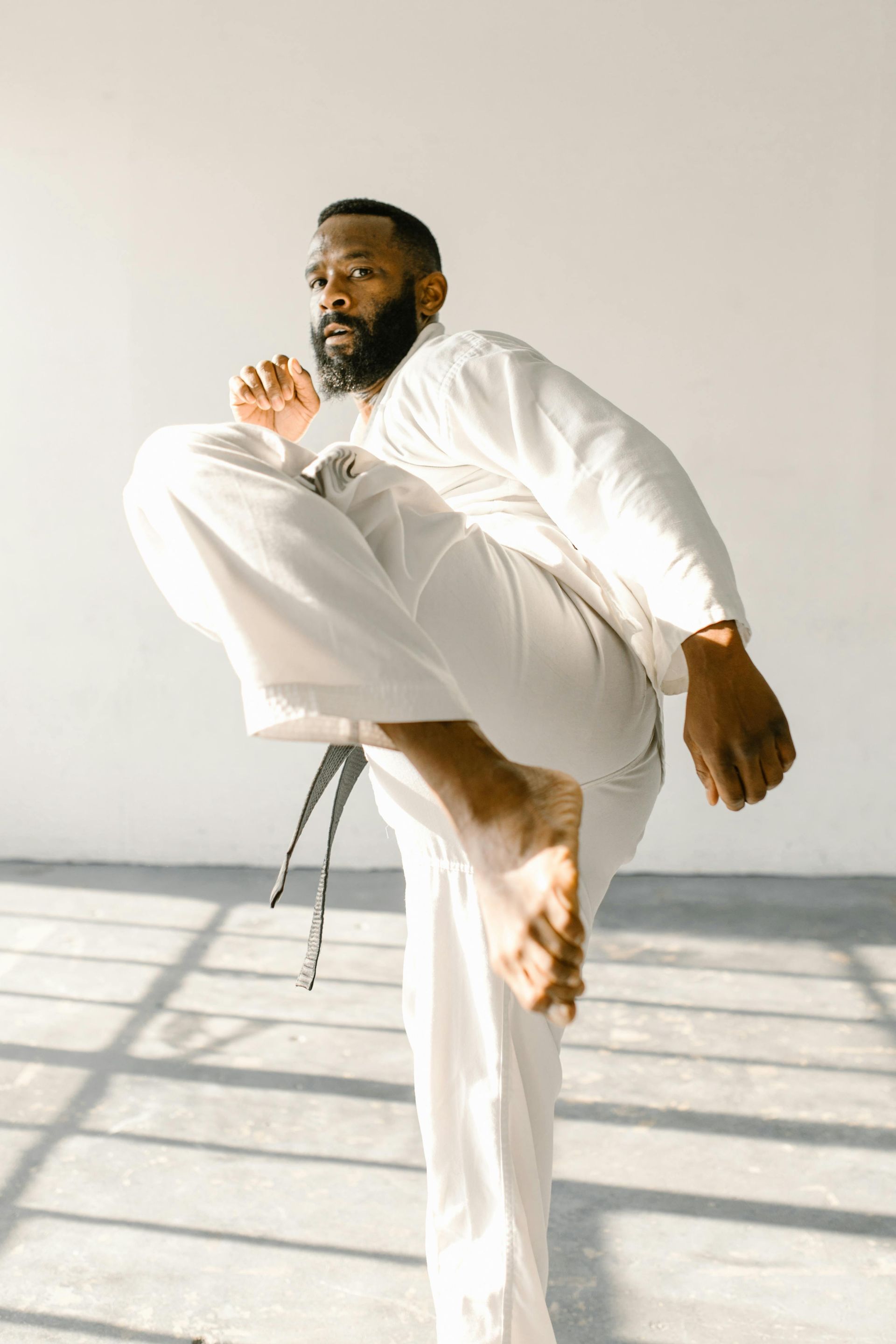 Man in white karate uniform kicking in a sunlit studio with a gray floor.
