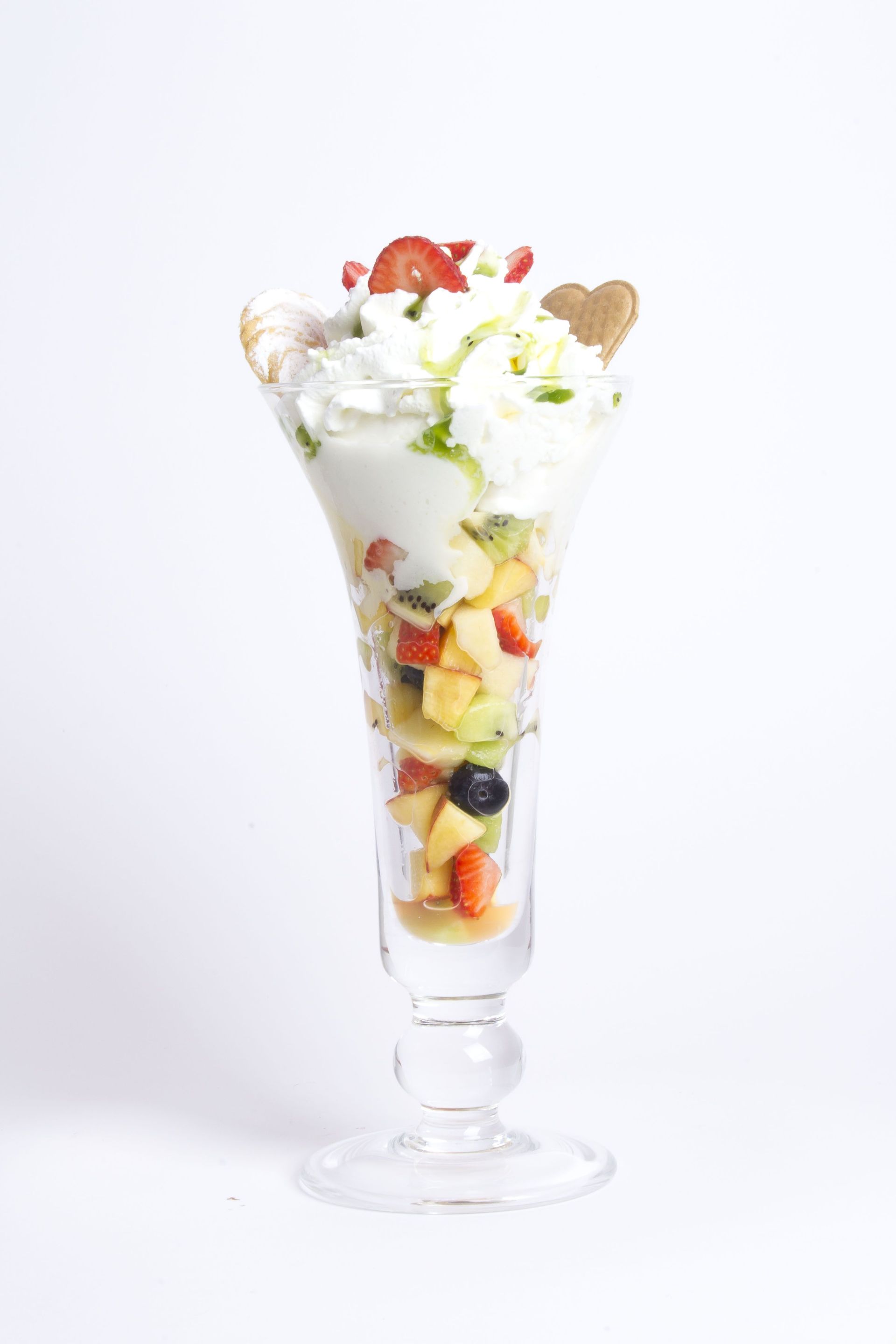 Un alto bicchiere da parfait colmo di strati di frutta fresca, panna e un biscotto a forma di cuore su uno sfondo bianco semplice.