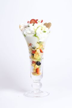 Un alto bicchiere da parfait colmo di strati di frutta fresca, panna e un biscotto a forma di cuore su uno sfondo bianco semplice.