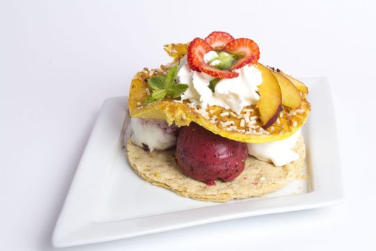 Un dessert elaborato con gelato, frutta, panna montata e sottili strati di pasta sfoglia su un piatto quadrato bianco.