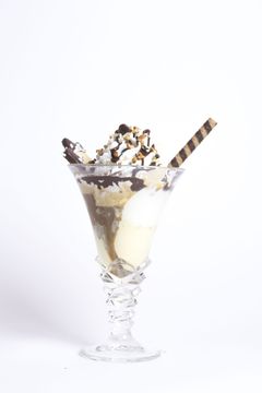 Una coppa di gelato in un bicchiere di cristallo, guarnita con panna montata, salsa al cioccolato, granella colorata e una cialda a righe, su uno sfondo bianco.