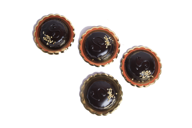 Quattro crostatine rotonde al cioccolato con bordi smerlati, ricoperte di ganache fondente e decorate con piccole foglie d'oro.