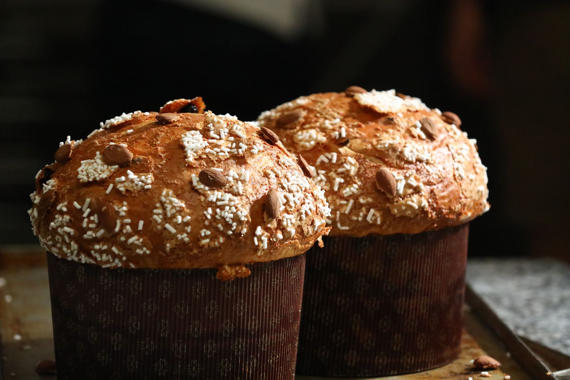 Due pagnotte di panettone guarnite con mandorle e granella di zucchero, adagiate su una teglia.