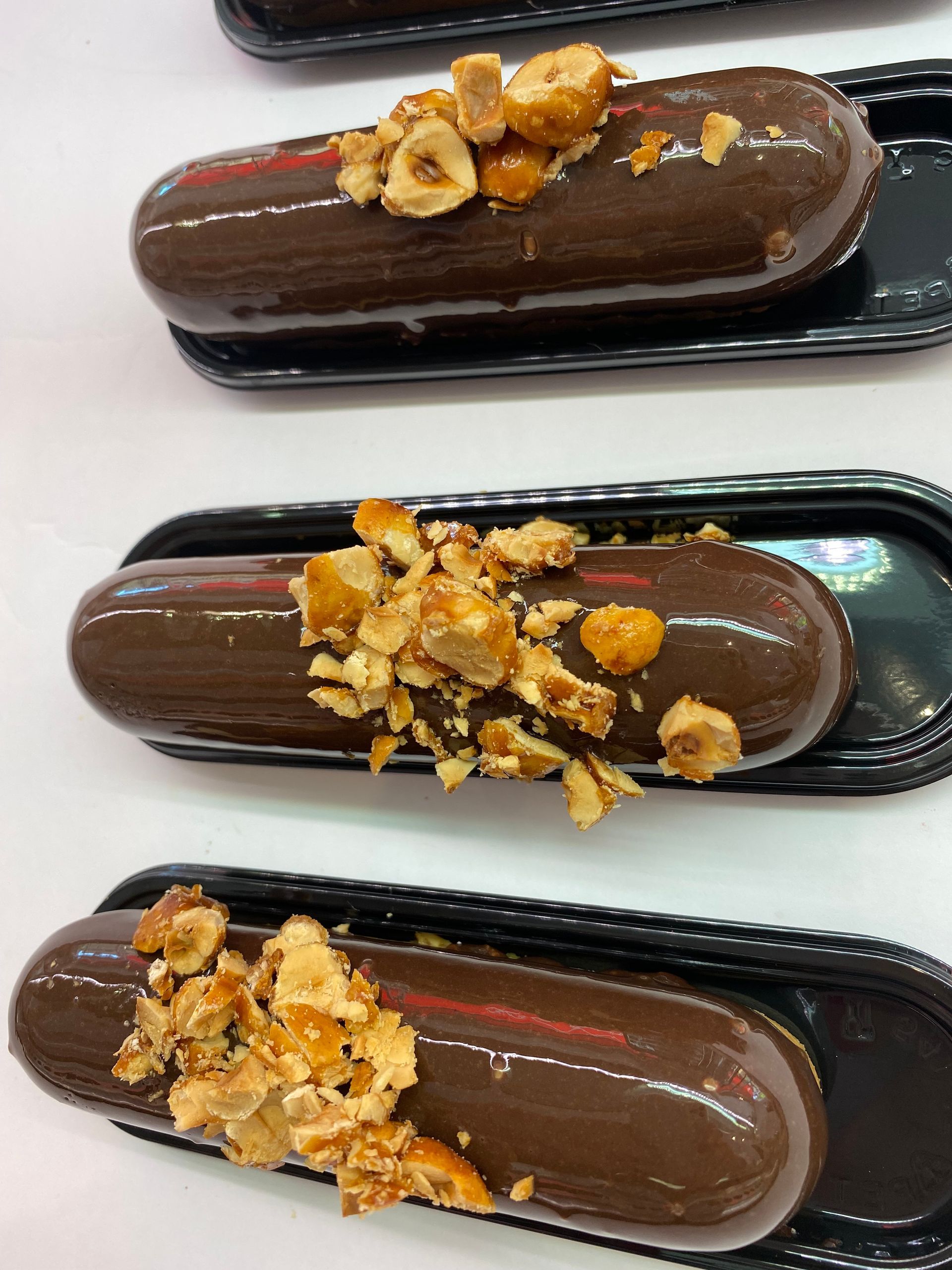Tre éclair ricoperti di glassa al cioccolato e guarniti con nocciole tritate, disposti in vassoi neri individuali.