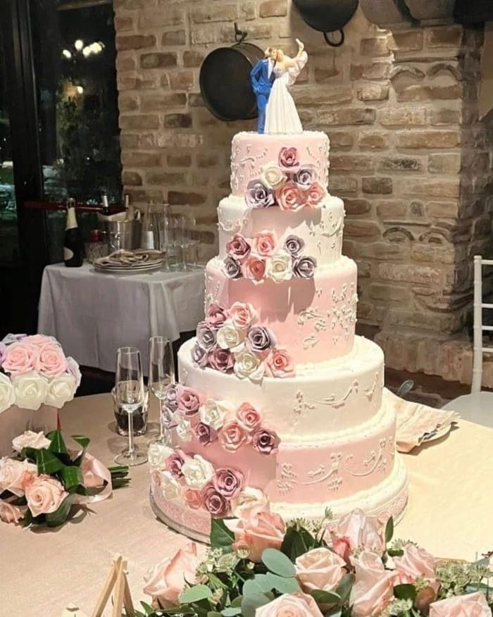 Una torta nuziale a cinque piani decorata con rose rosa e bianche, sormontata da una statuina degli sposi, in un ambiente rustico al chiuso.