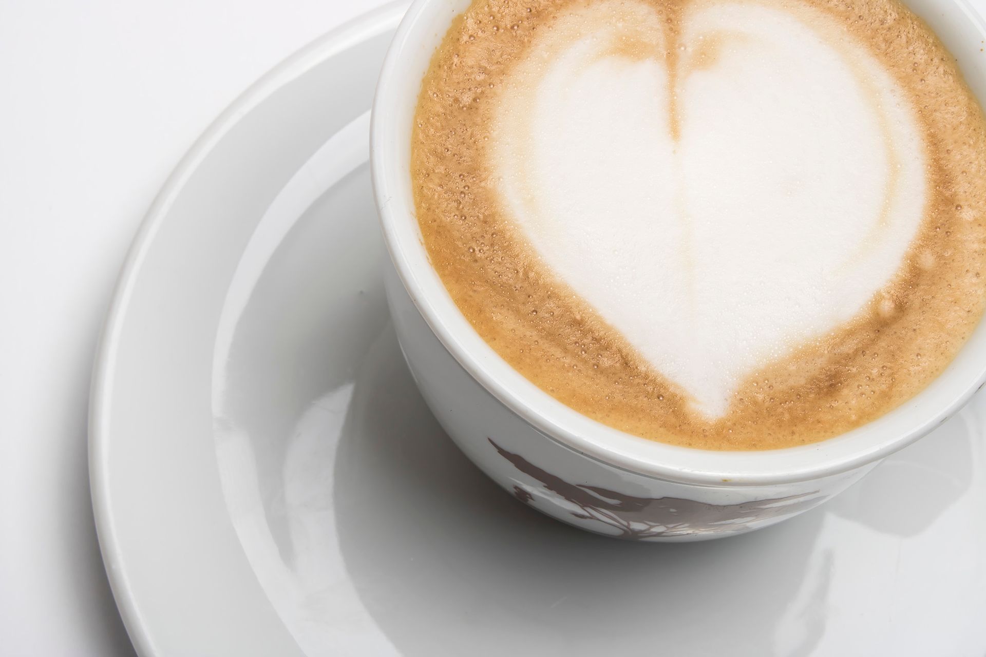 Una tazza bianca decorata con latte art a forma di cuore, servita su un piattino bianco abbinato su uno sfondo neutro.
