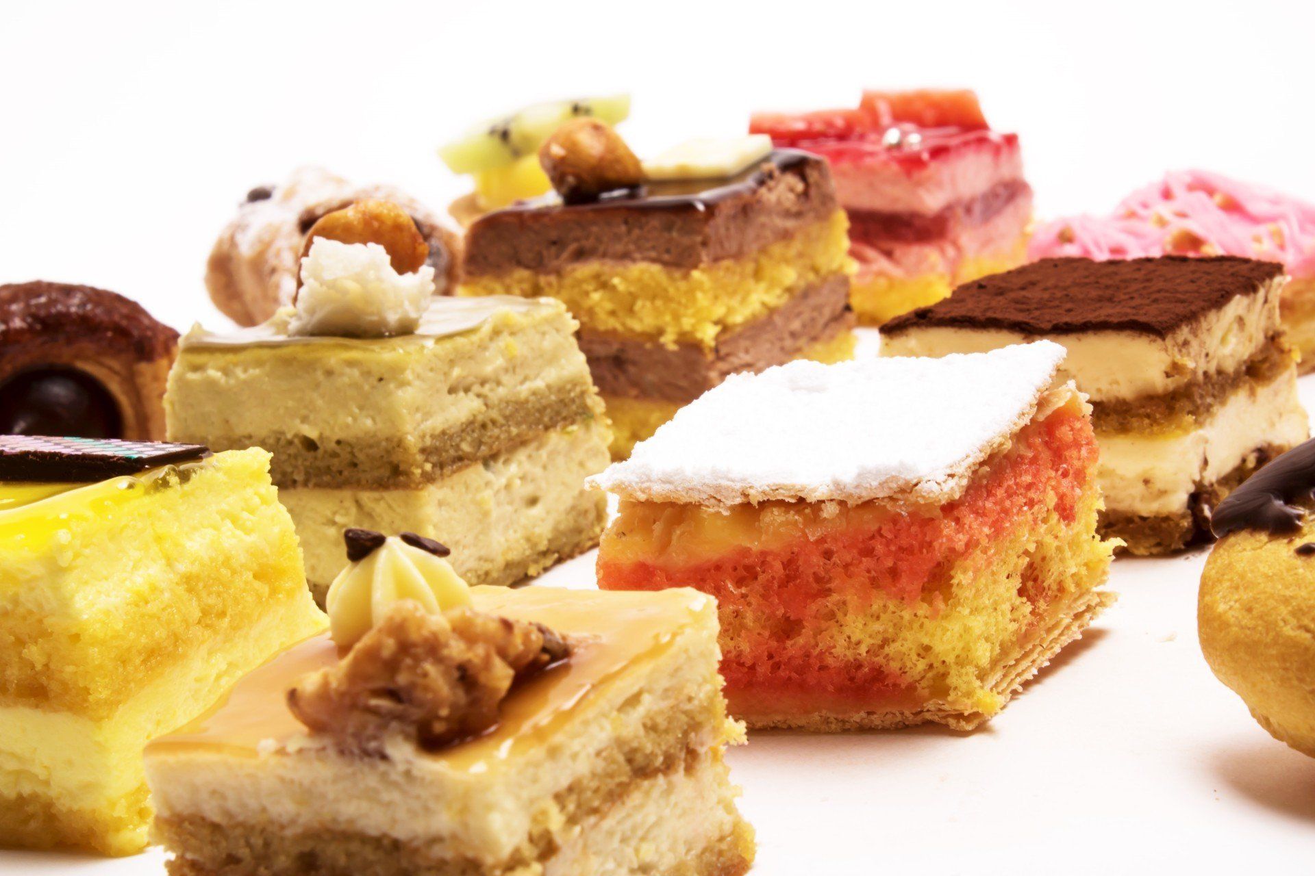 Un assortimento di piccole torte e pasticcini rettangolari di vario tipo, con guarnizioni colorate, disposti su uno sfondo bianco.