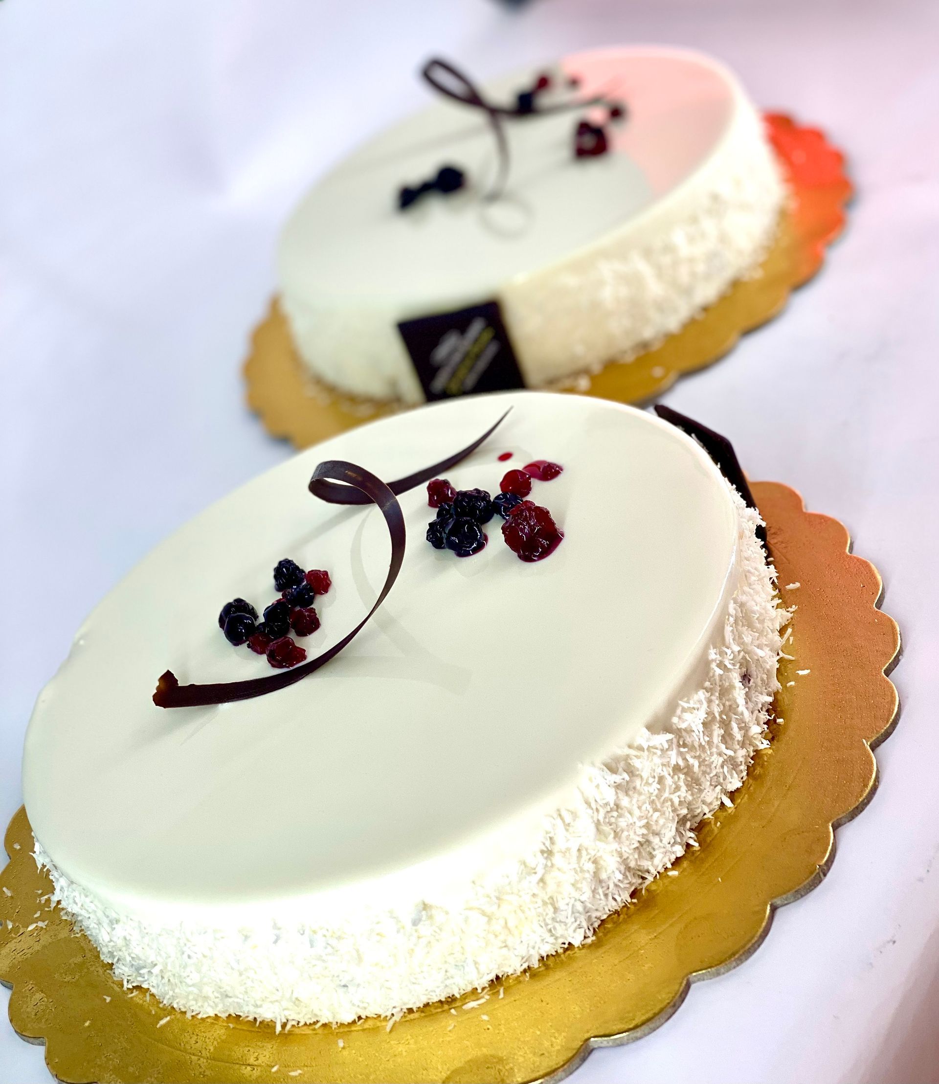 Due torte rotonde, ricoperte di glassa bianca e decorate con frutti di bosco e riccioli di cioccolato, su basi di cartone dorato.