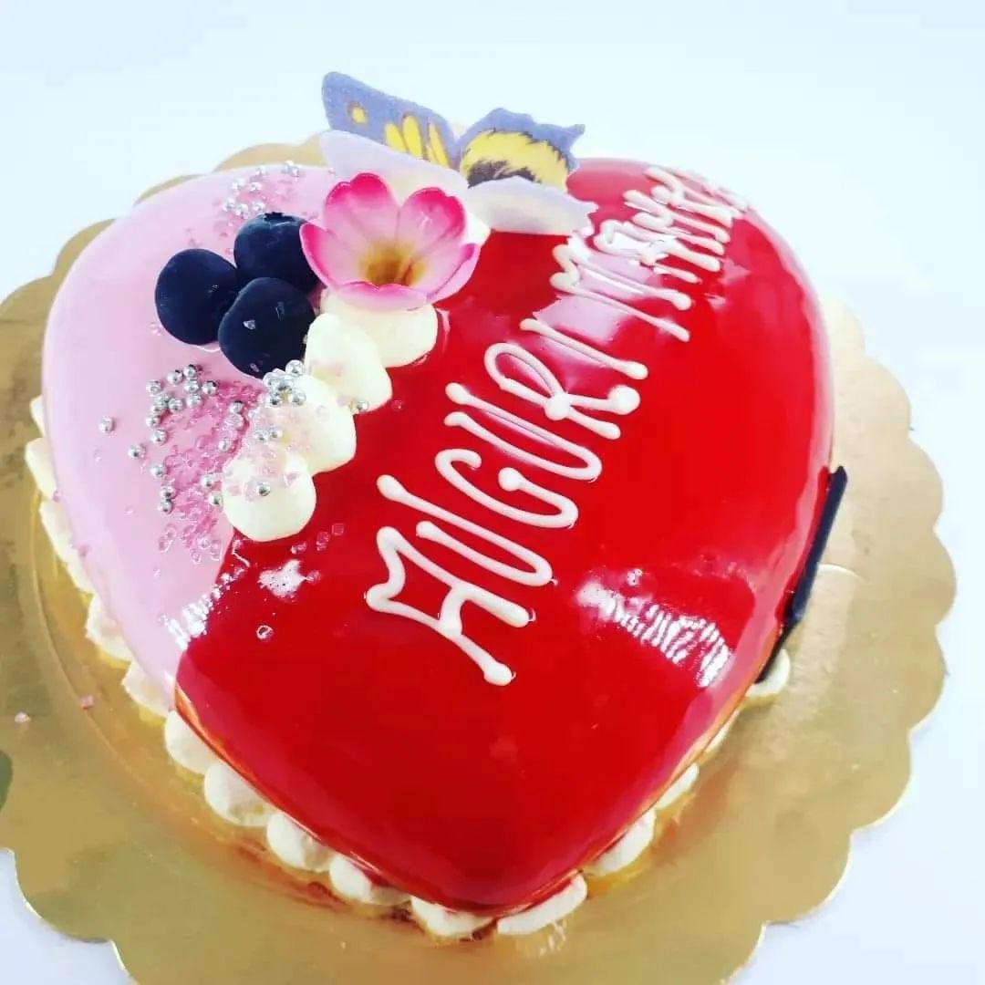 Una torta a forma di cuore con glassa rosa e rossa, decorata con mirtilli, un fiore, una farfalla e la scritta 