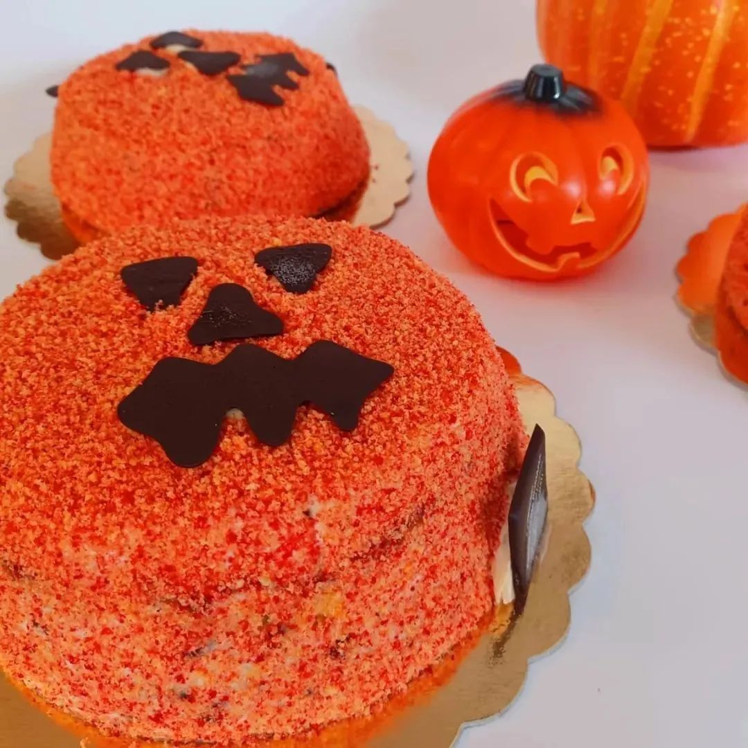 Due torte arancioni a forma di zucca, dalla consistenza particolare, con facce di zucca intagliate in cioccolato fondente, accanto a zucche decorative di plastica.