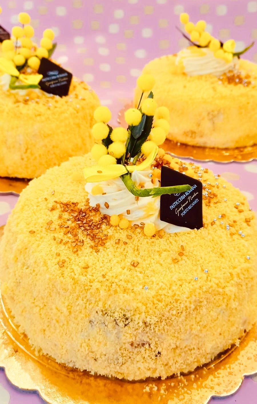 Tortine al gusto mimosa gialla decorate con piccole rosette di panna montata e rametti di fiori gialli su basi per torte dorate con bordi smerlati.