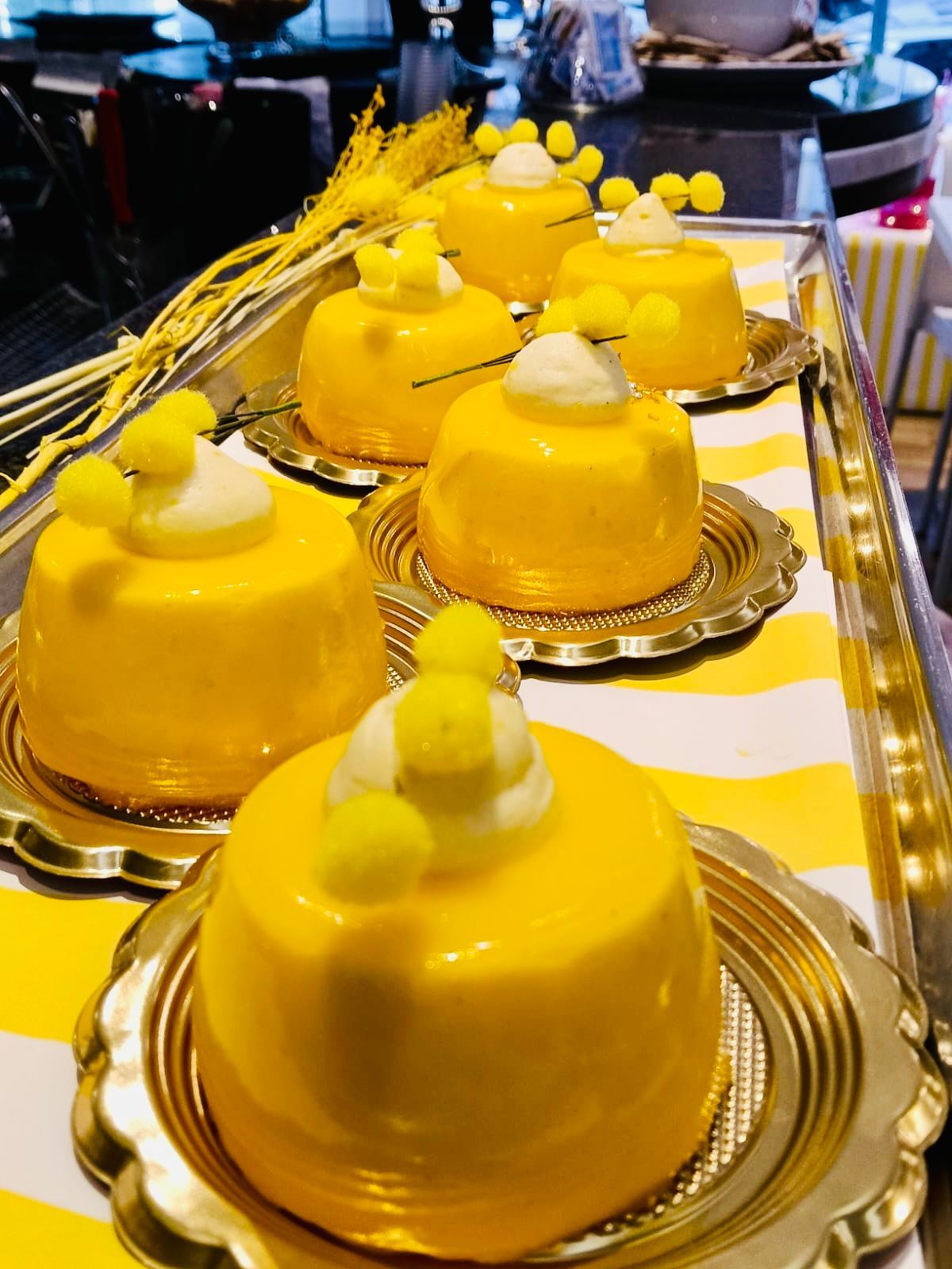 Diverse mini torte glassate di un giallo brillante, decorate con scorza d'arancia candita, servite su piatti dorati smerlati e guarnite con mimosa.