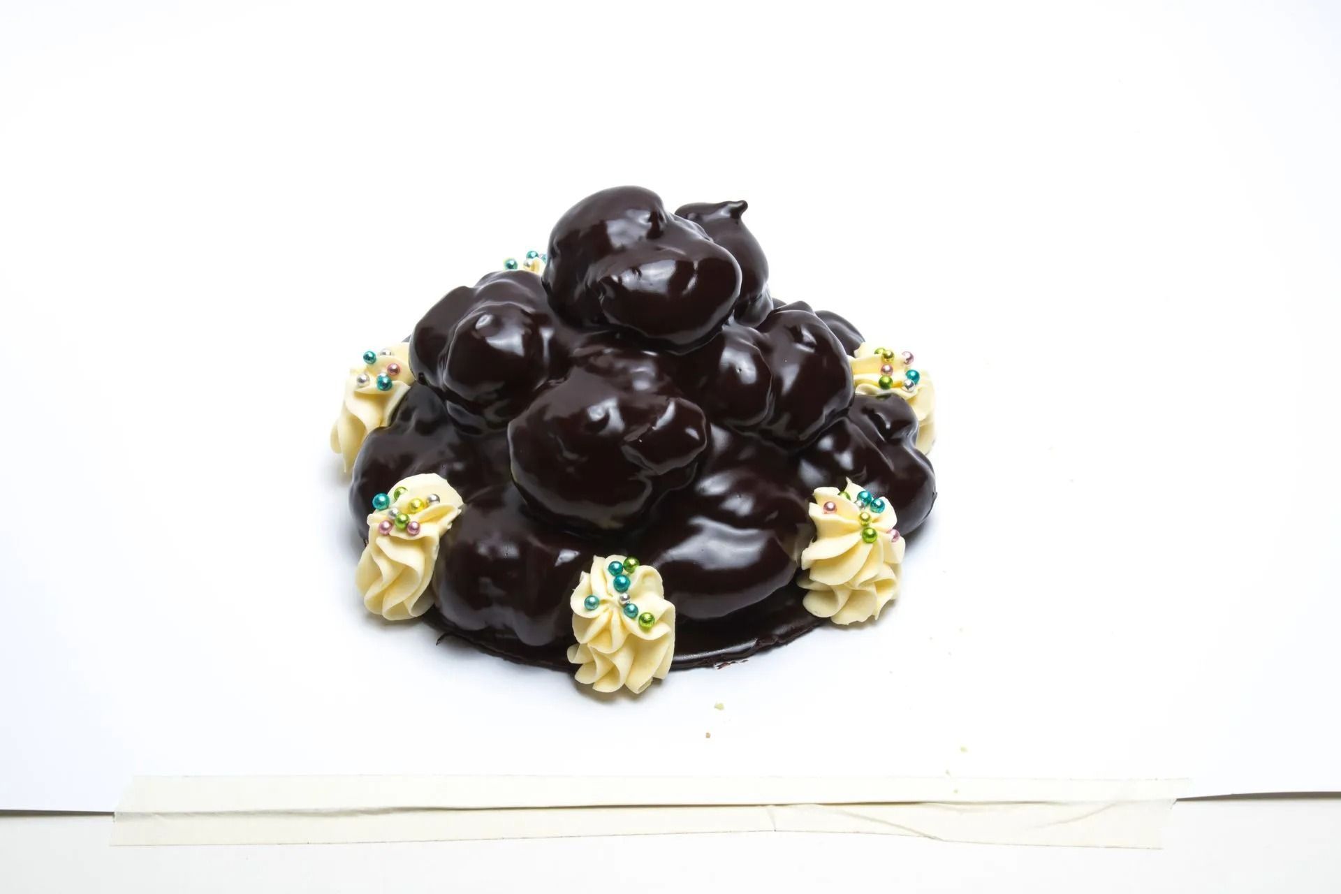 Un dessert a forma di torre di profiteroles al cioccolato, guarnito con ciuffi di panna montata e minuscole codette blu su sfondo bianco.