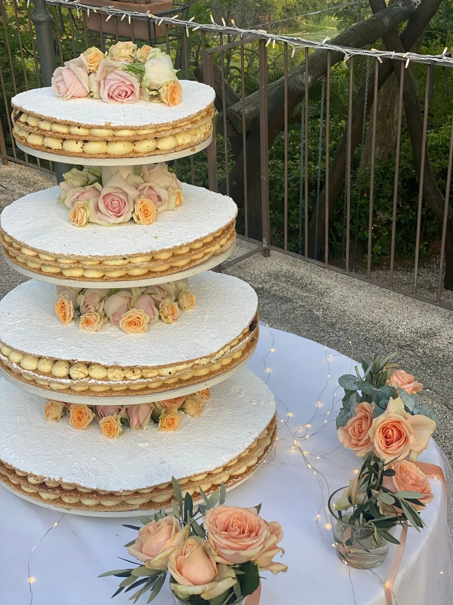 Una torta nuziale a quattro piani composta da macaron e decorata con pesche fresche e rose rosa, posizionata su un tavolo all'aperto.