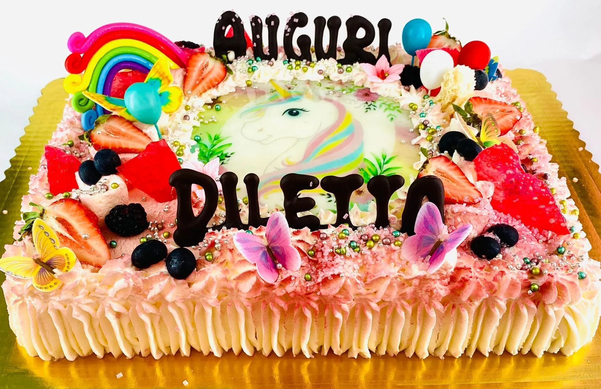 Una torta di compleanno rettangolare decorata con un unicorno, un arcobaleno, fragole, frutti di bosco e farfalle, con la scritta 