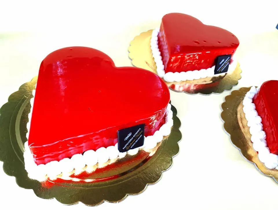 Tre torte a forma di cuore con glassa rossa lucida e decorazioni di crema bianca, adagiate su vassoi dorati con bordi smerlati.